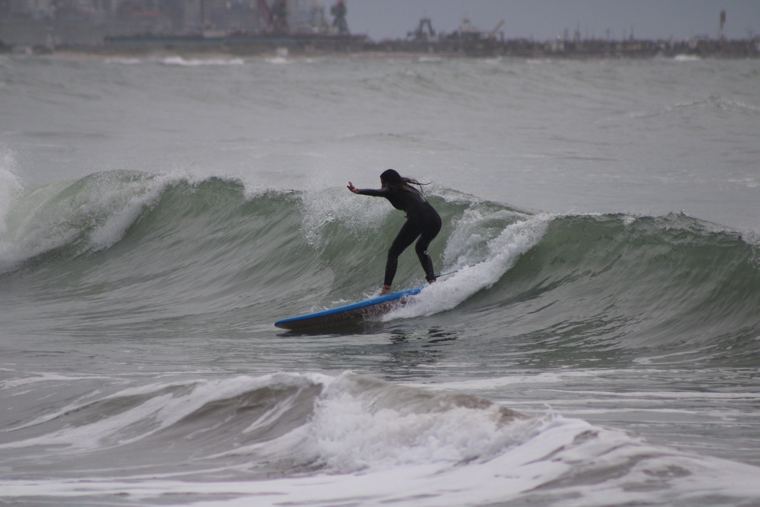 Alumna surfeando una ola con el puerto de fondo